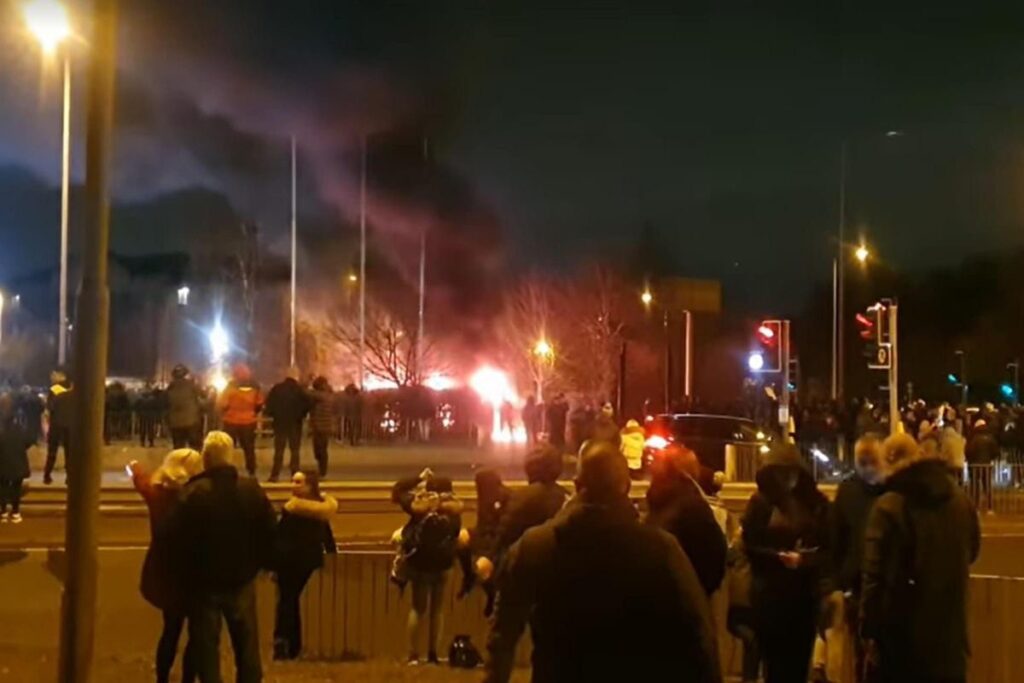 A crowd are standing and watching by the side of a road in Knowsley. In the background smoke and fire can be seen from the riot.