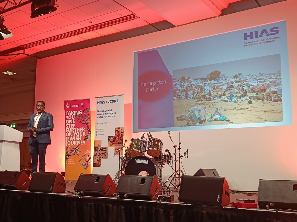 Monim Haroon presents on stage at Limmud. He is wearing a suit, and behind him are Limmud and HIAS+JCORE banner stands. Projected behind him is a screen showing a slide, with an image of a refugee camp and text reading: "The forgotten Darfur".