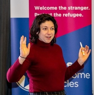 Meiron Avidan is presenting at an event. She is stood in front of a HIAS banner stand advertising Welcome Circles. She has her hands raised to make a point.