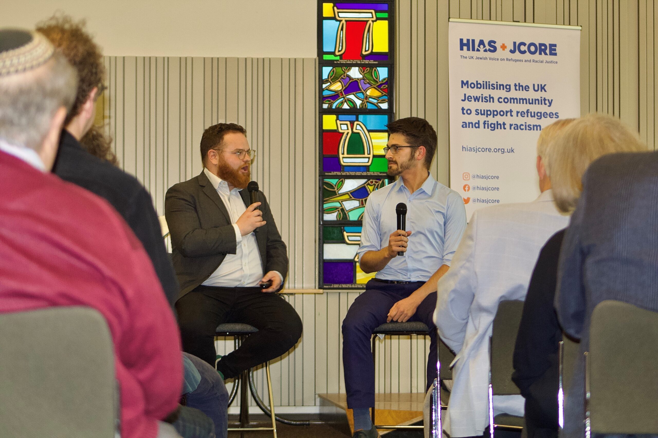 Image is from behind two sections of the crowd on the left and the right. In the middle, Amos Schonfield (HIAS+JCORE's Deputy Director) and Josh Stein (a JUMP volunteer) can be seen talking on stage. They are seated and both holding microphones.