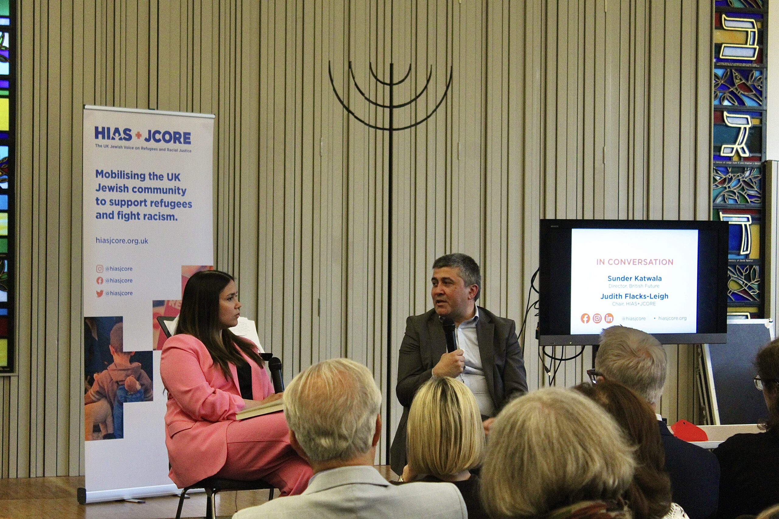 Judith Flacks (HIAS+JCORE's Chair) and Sunder Katwala (British Future Executive Director) are speaking on stage and sat down in chairs. They are in front of an audience. Sunder is sat on the right and speaking into his microphone. Judith is sat on the left and holding a microphone on her lap. Behind them is a screen displaying 