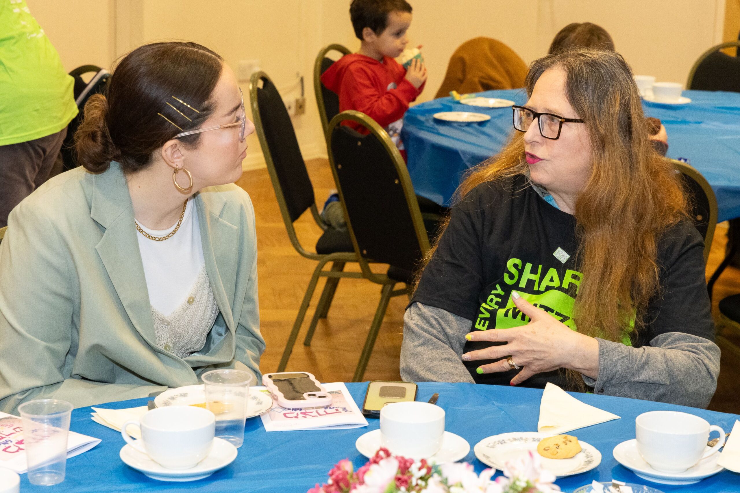 Photo shows Eliza Ward, HIAS+JCORE's Frontline Support Manager, talking to a woman wearing a Mitzvah Day t-shirt. Eliza is wearing a grey suit jacket and both are seated at a table.