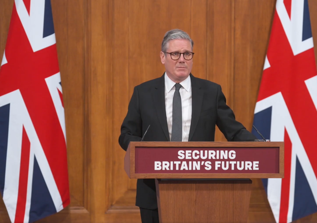 Sir Keir Starmer stands at a lectern. To his left and right are large Union Jack flags.